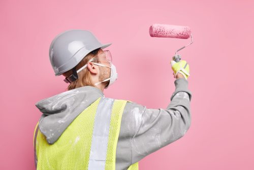 Sideways shot of busy handyman dressed in safety uniform holds paint roller wears protective mask works hard isolated over pink background. Repair and renovation concept. Maintenance worker.